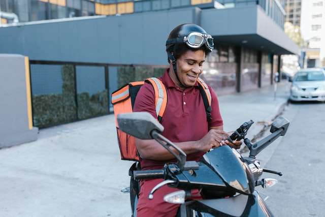 Foto de um rapaz em cima de uma moto com o celular na mão sorrindo e olhando para o celular